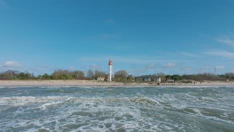 Aerial-establishing-view-of-white-colored-Pape-lighthouse,-Baltic-sea-coastline,-Latvia,-white-sand-beach,-large-waves-crashing,-sunny-day-with-clouds,-low-drone-orbit-shot