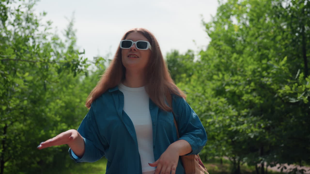 선글라스와 흰색 의상에 파란색 셔츠를 입고 푸른 식물에 둘러싸여 기쁘게 춤을 추는 젊은 여성, 공원 길에서 여름 햇살을 즐기며 움직임 속에서 미소 짓고 있어요.