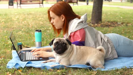 chica acostada y escribiendo en una computadora portátil en un césped con su pug alrededor