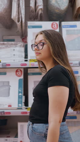 mujer de compras en una tienda minorista caminando más allá de un estante lleno de productos de cama envasados, mira a su alrededor mientras explora opciones, pasillo de la tienda bien iluminado lleno de artículos para el hogar y exhibiciones organizadas