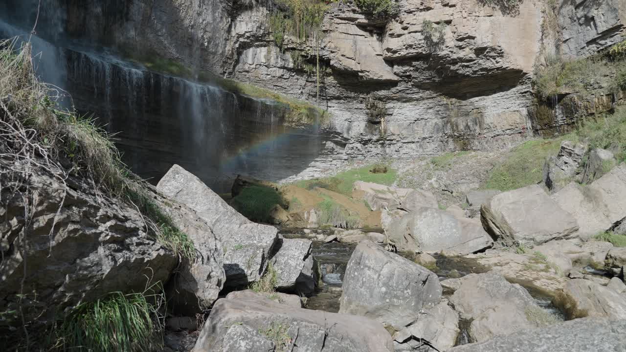 A serene waterfall cascades over rocky cliffs with a faint rainbow on a sunny day