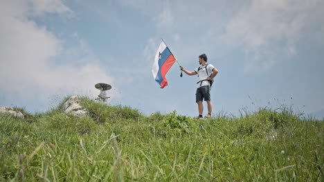 Kamerafahrt-Aus-Niedriger-Perspektive-Einen-Hügel-Hinauf-Ein-Wanderer-Oben-Mit-Einem-Wanderstock-In-Der-Hand-Und-Einer-Daran-Befestigten-Slowenischen-Flagge,-Die-Sie-Im-Wind-Flattern-Lässt