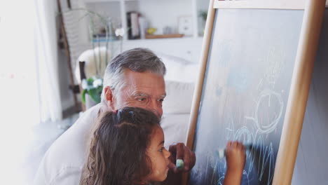 Senior-man-drawing-on-a-blackboard-with-his-young-granddaughter-at-home