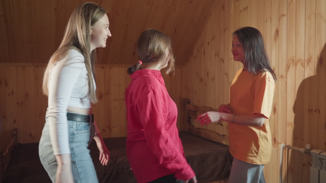 three female friends enter cozy wooden room smiling and interacting before relaxing together on brown bed, casual indoor moment with visible fan in corner