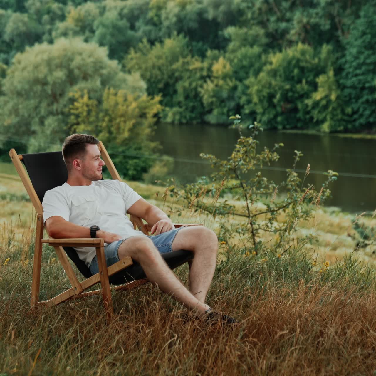 Bearded Caucasian man in white t-shirt sits in a folding chair. Happy relaxed man smiles enjoying the nature landscape