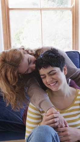happy caucasian lesbian couple sitting,embracing,kissing and smiling in sunny living room