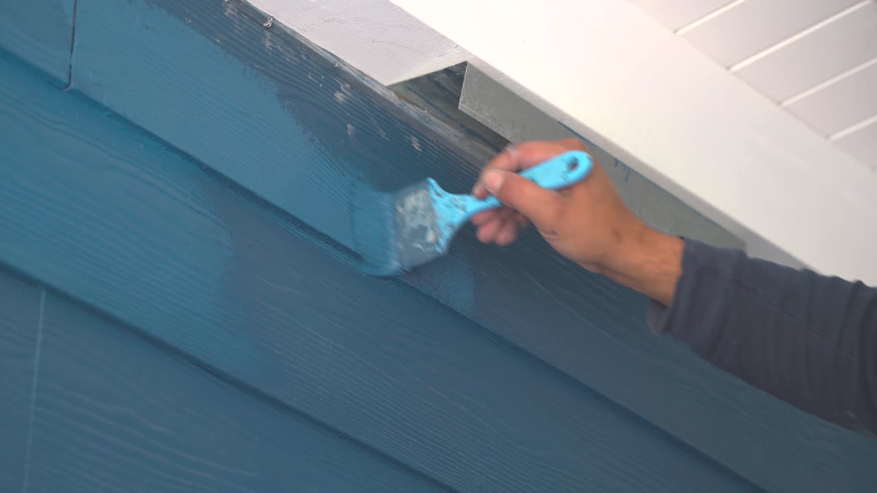 Gros plan sur la main d'un peintre peignant en bleu la surface d'un mur en bois en extérieur par une journée ensoleillée.
