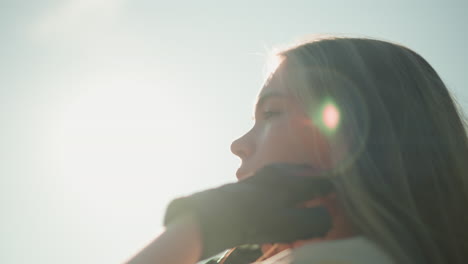 white lady in black gloves tilts her head back, adjusting her hair with sunlight creating a glowing effect around her face, softly blurred greenery in the background