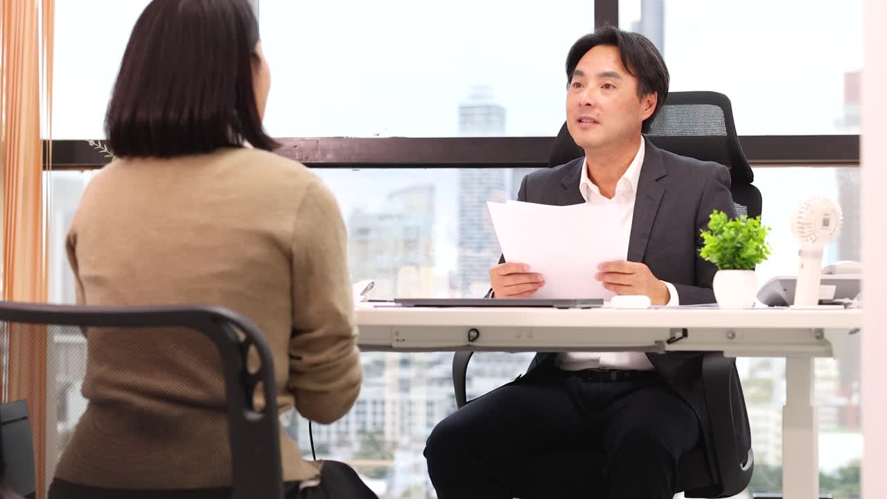 Professional Asian recruiter reviews documents while interviewing a candidate in a bright city office