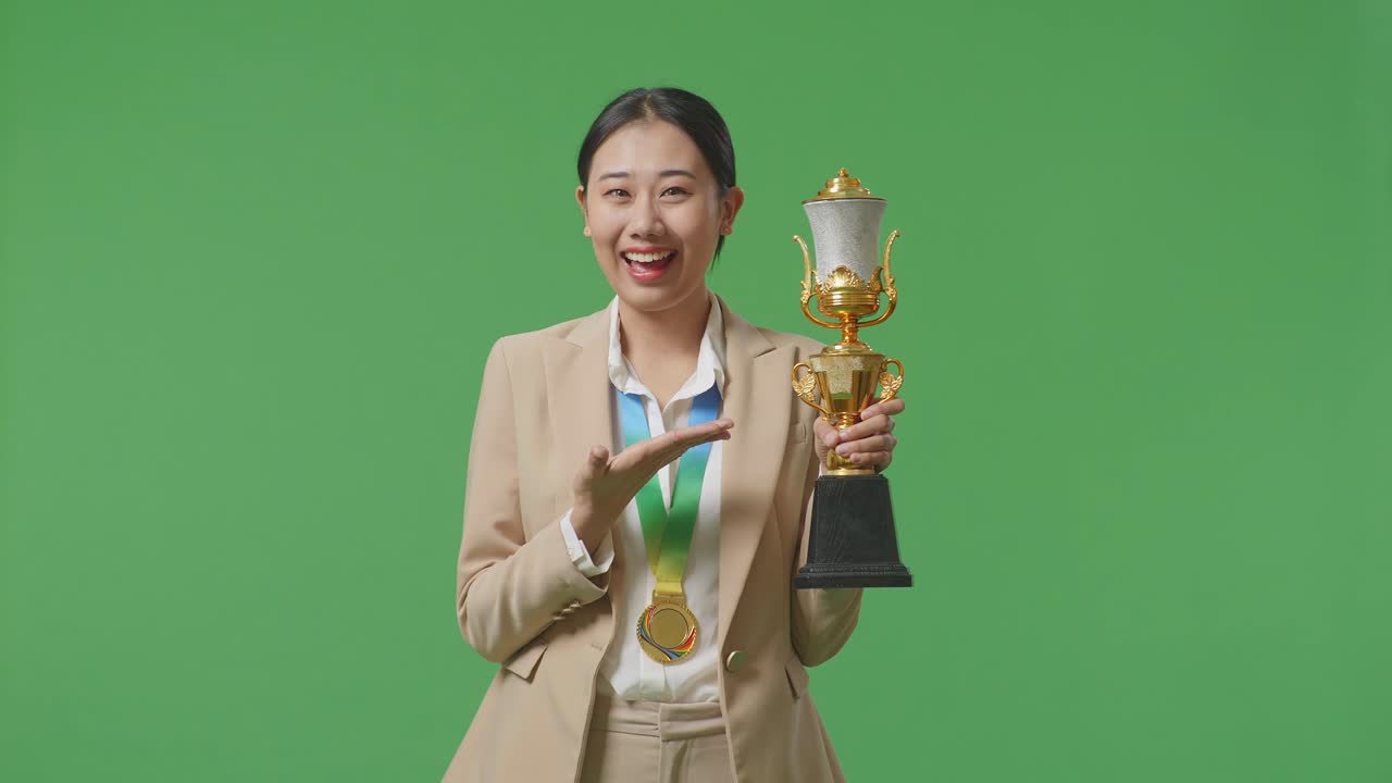 금메달을  슈트를 입은 아시아 사업가 여성은 손에 금 트로피를 가리키며 미소 짓고 스튜디오의 녹색 화면 배경에서 첫 번째 우승자로 자랑스럽게 승리합니다.