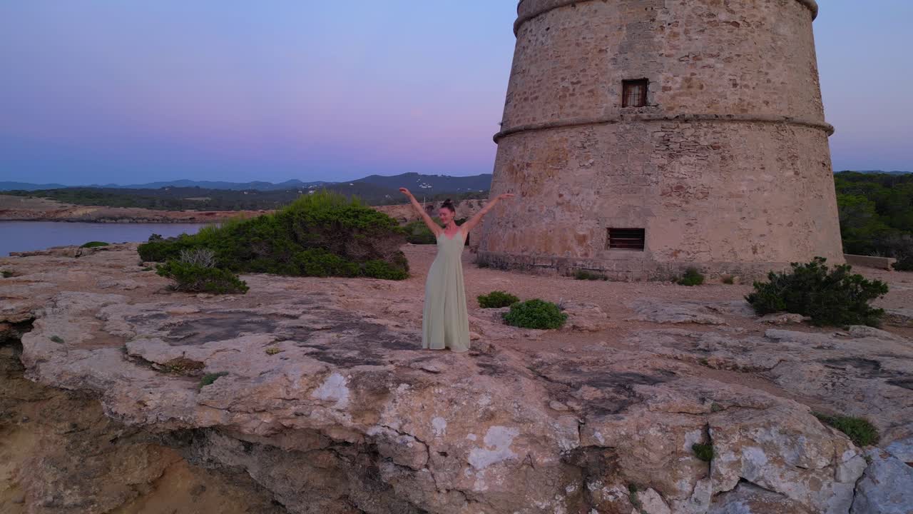Barefoot hippie girl practicing yoga at sunset near Torre des Carregador, Ibiza, Spain. Fantastic aerial view flight static tripod hovering drone