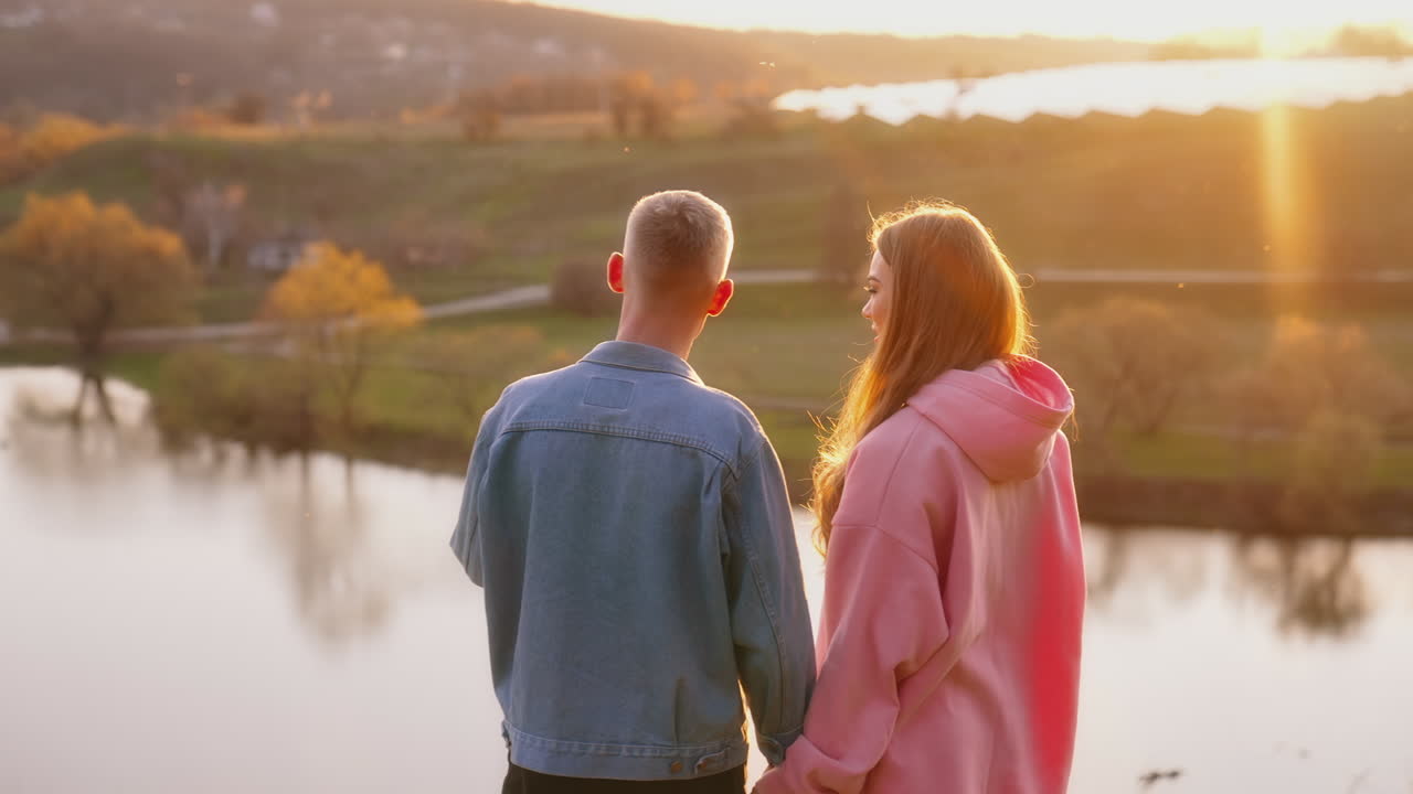 Couple enjoys beautiful evening landscape in the countryside. Young man and woman standing on the hill and looking on the river at sunset in autumn time.