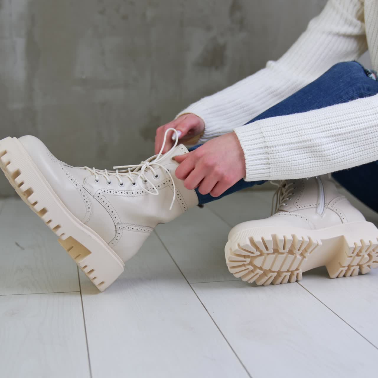 Female model wearing white sweater and jeans sits on the floor. Lady touching her stylish white boots on tractor soles. Close up