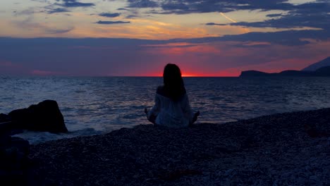 Yoga-Pose-Eines-Mädchens,-Das-Auf-Kieselsteinen-Eines-Ruhigen-Strandes-Sitzt,-Der-Vom-Meer-Umspült-Wird-Und-Den-Farbenfrohen-Sonnenuntergang-Widerspiegelt