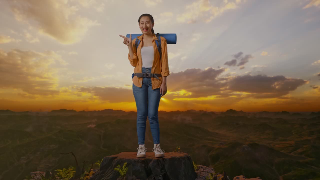 cuerpo lleno de excursionista femenina asiática con mochila de montañismo sonriendo y apuntando a un lado mientras está de pie en la cima de la montaña durante la hora del atardecer