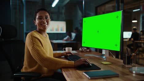 joven especialista multiétnico trabajando en una computadora de escritorio con pantalla verde simulacro de visualización en la oficina creativa. hermosa gerente femenina diversa con cabello corto y gafas está usando un jumper amarillo.