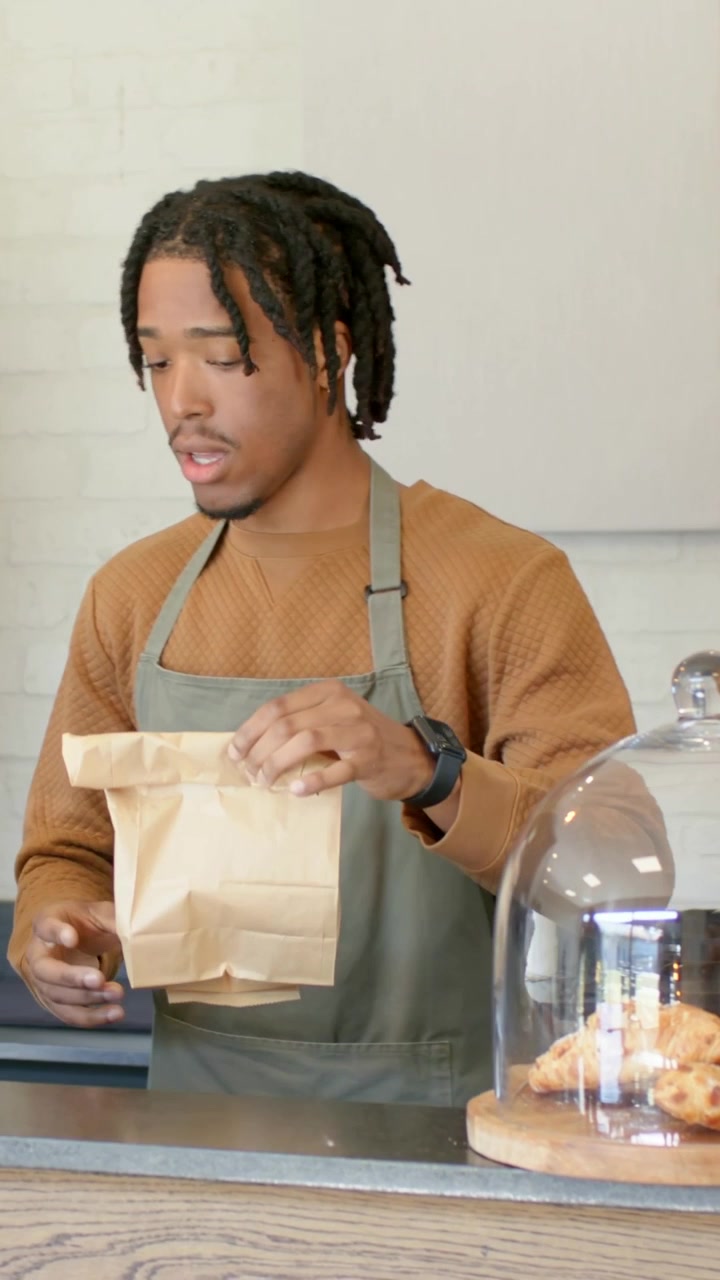 Vertical video: Order coming, barista in green apron folding brown paper bag for pickup at counter