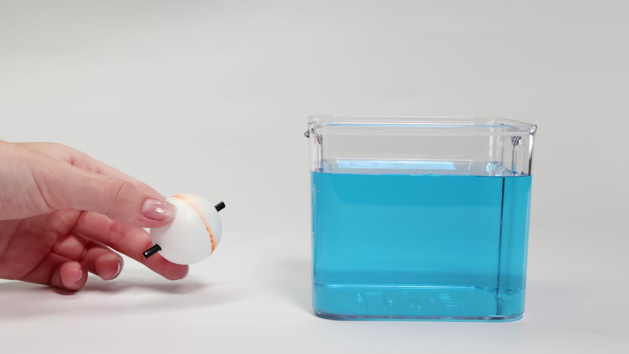 A hand drops a foam ball into blue liquid, demonstrating buoyancy. The ball floats, showcasing principles of density and flotation