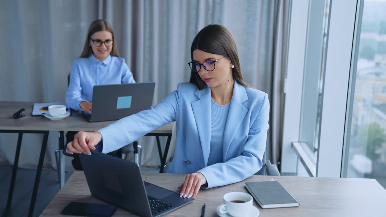 Two long-haired women wearing glasses sitting at desks in the office. Dark-haired lady opens her laptop and then paper notebook and takes a pen. Female colleague working at the computer at backdrop.
