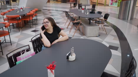 elegant woman seated in a mall engaged in a phone call, hand resting on the table in a cafe, with chairs arranged in the background and a partial view of other shoppers passing by