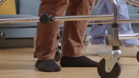 cerca de los pies del paciente masculino caminando por un andador al lado de una enfermera que lo ayuda durante una terapia física en casa