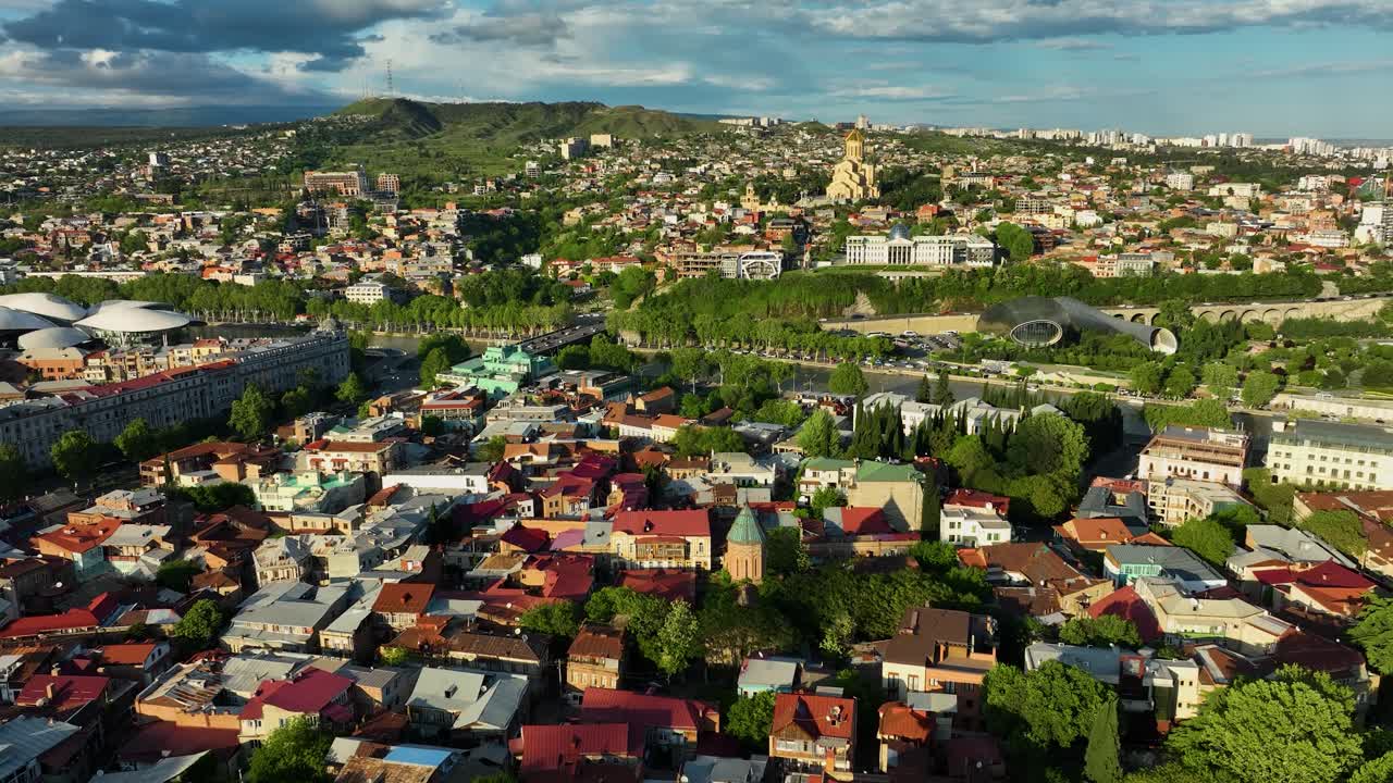 Wide aerial shot showing Tbilisi’s old town with the Holy Trinity Cathedral rising above colorful buildings and green hills
