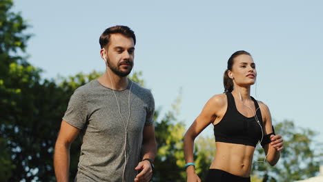 Vista-De-Cerca-De-Una-Pareja-De-Jóvenes-Corredores-Corriendo-Juntos-Escuchando-Música-Con-Auriculares-En-El-Estadio-En-Una-Mañana-De-Verano