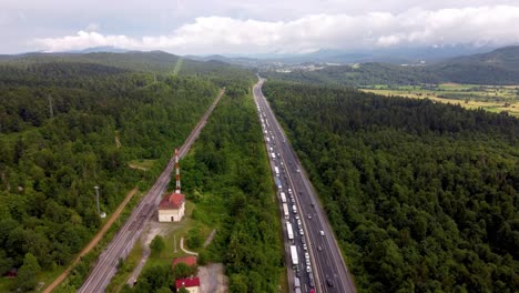 autopista europea con tráfico pesado y congestión y líneas ferroviarias y eléctricas que forman tres capas de líneas orientadas en la misma dirección con nubes y colinas en el fondo con cinturón verde de árboles