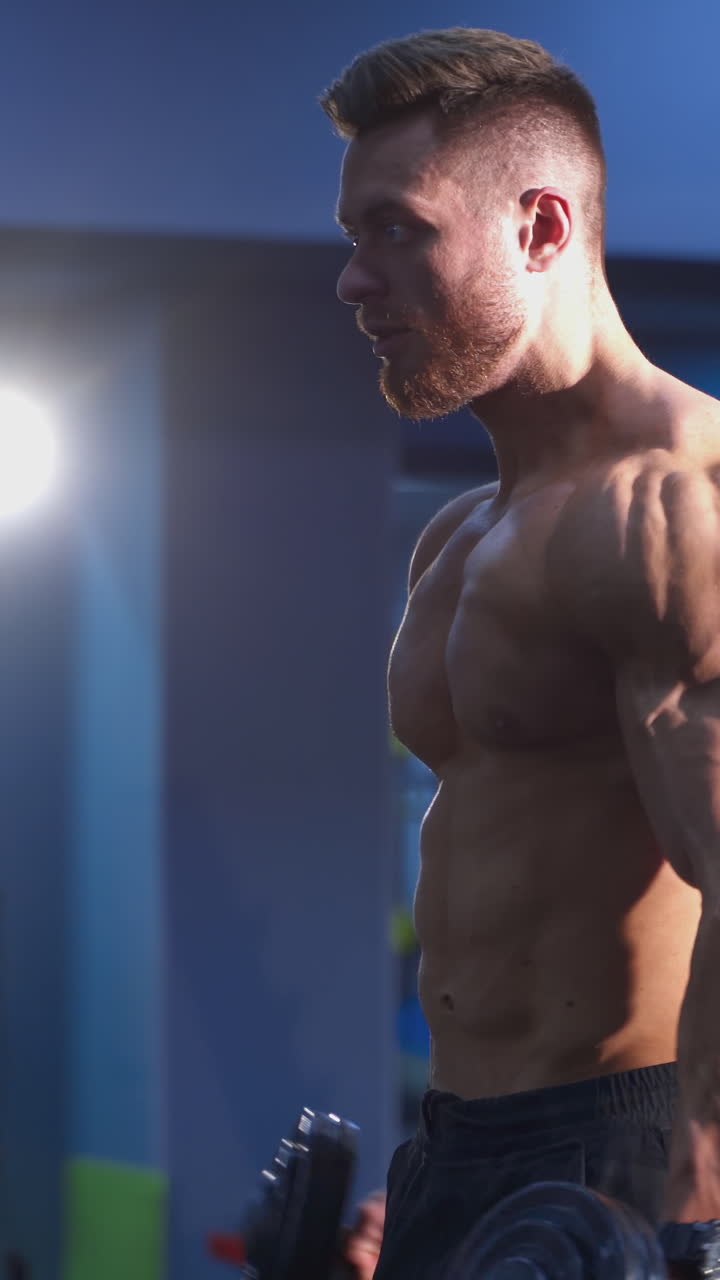 Athlete with heavy dumbbells training indoors. Strong man with muscular naked body is lifting weights in fitness center. Daily workout of a bodybuilder. Vertical video