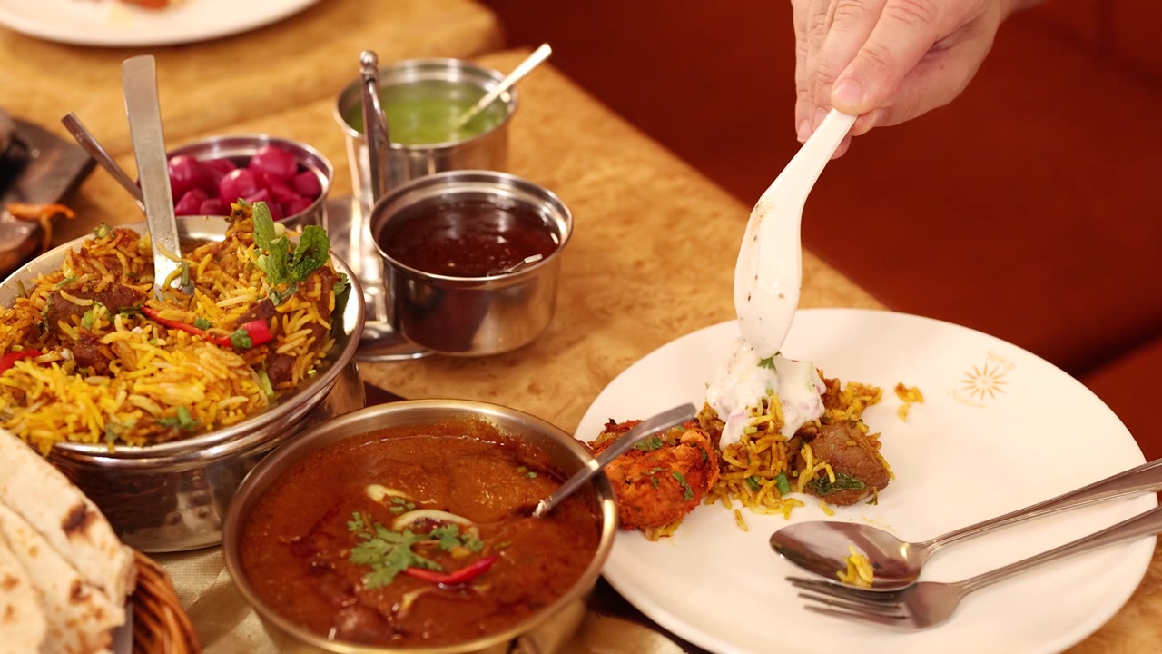 En hånd hælder hvid chutney over biryani-ris blandt et udvalg af indiske retter.