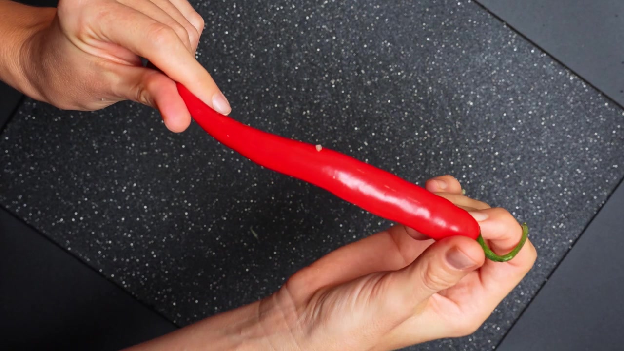 Cooking with red chili in a kitchen setting during the day. Person holds a long red chili in their hands while preparing ingredients in a kitchen.