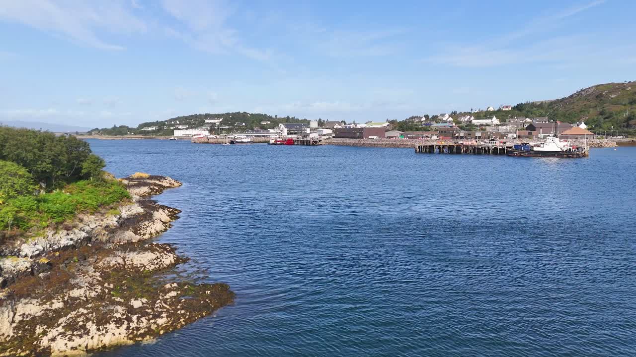 Aerial drone view over rocky coastline toward harbor, boats, and village beneath a clear sky