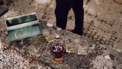 un hombre sirviendo whisky en vasos en una rústica mesa de piedra al aire libre.