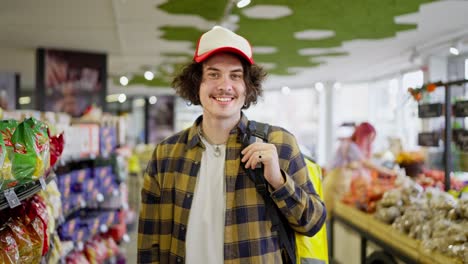 Portrait-of-a-Happy-food-delivery-guy-with-curly-hair-in-a-plaid-shirt-who-stands-in-the-middle-of-a-supermarket-while-choosing-products-for-delivery