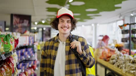 retrato de un tipo de entrega de comida feliz con cabello rizado en una camisa a cuadros que se encuentra en el medio de un supermercado mientras elige productos para la entrega