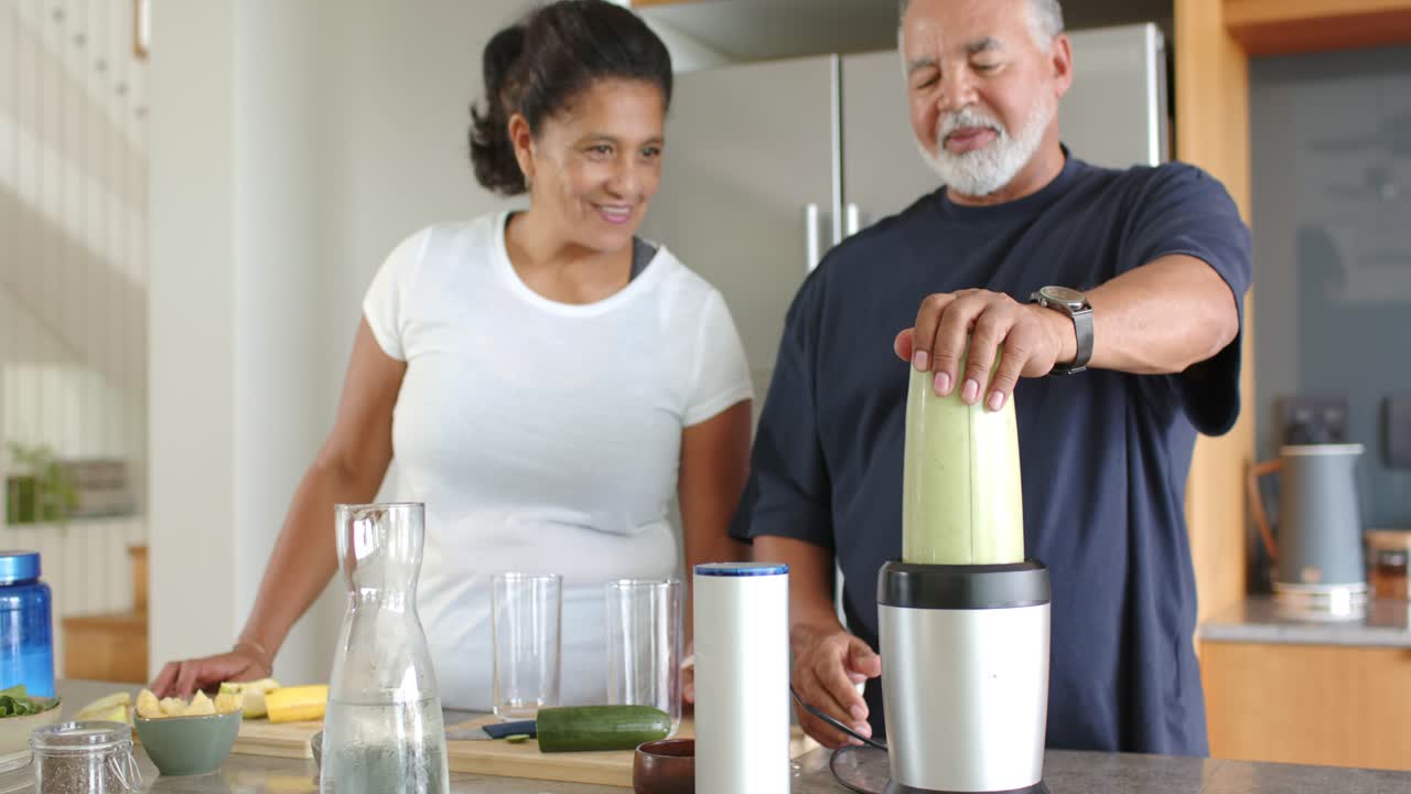 Coppia anziana che posiziona la tazza del frullatore sulla base sull'isola della cucina, frullando e preparando un frullato verde.