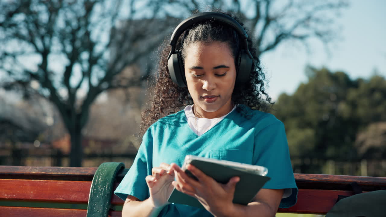 Woman in scrubs using a tablet with headphones in the park