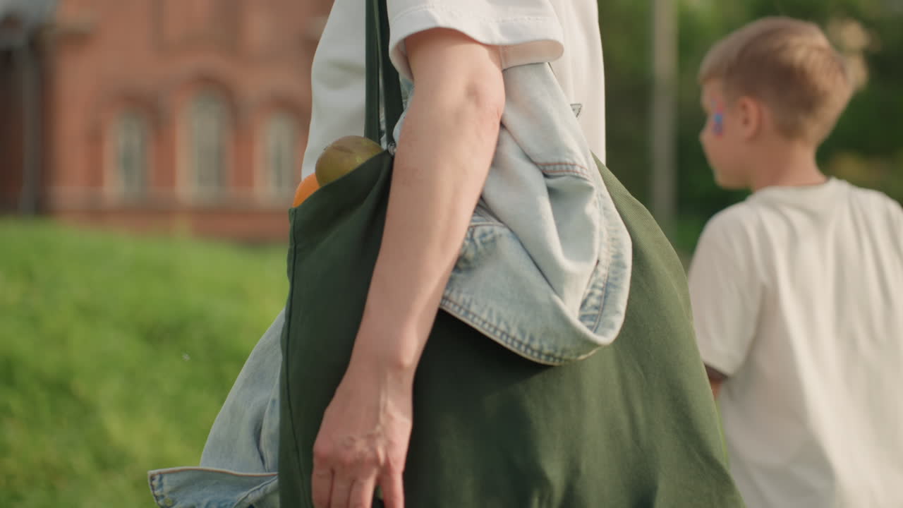 Back view of housewives walking with kids in sunny park holding hands, blurred building in distance, casual summer clothing, family outing stroll conveying relaxed afternoon connection
