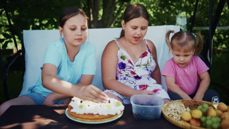 three girls enjoying dessert outdoors with cake, fruits, and a sunny garden setting