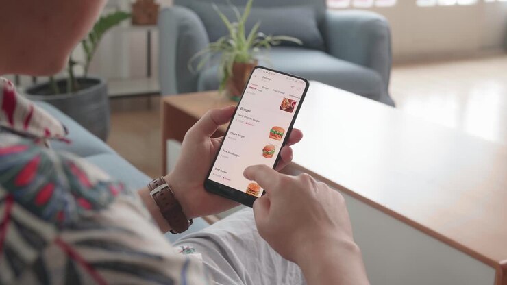 Man Sitting On Couch Using Smartphone Buys In Internet Shop. Male Selects Food In Online Store