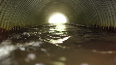 Flood-water-running-through-corrugated-pipe-with-light-at-end,-zoom-shot