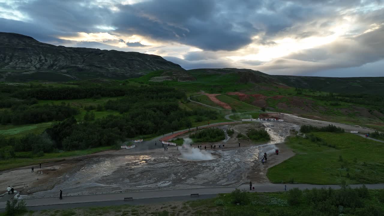 Drone shot of an eruption at Geysir geothermal area in Iceland, surrounded by green hills, dramatic skies, and mountain views at sunset.