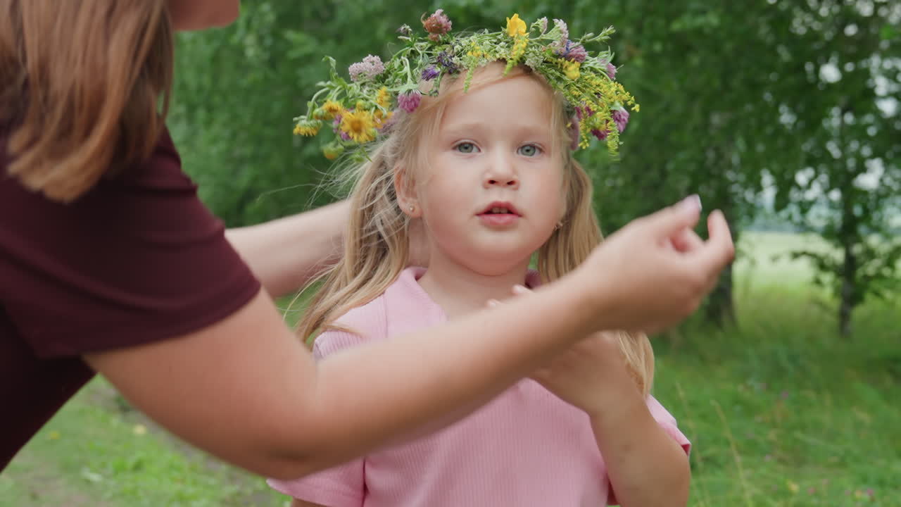 Kleines kaukasisches Mädchen mit Blumenkranz, Mutter richtet den Kranz und gibt sanfte Küsse, grüner Birkenhintergrund, weiches Sommerlicht, zärtliche Familienbande und kindliche Geborgenheit, offenes Porträt