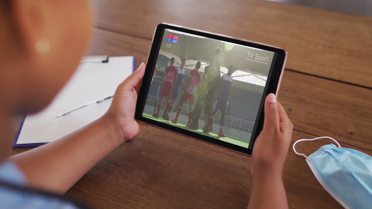 태블릿에서 하키 경기를 시청하는 얼굴 마스크와 함께 테이블에 앉아있는 여성의 복합