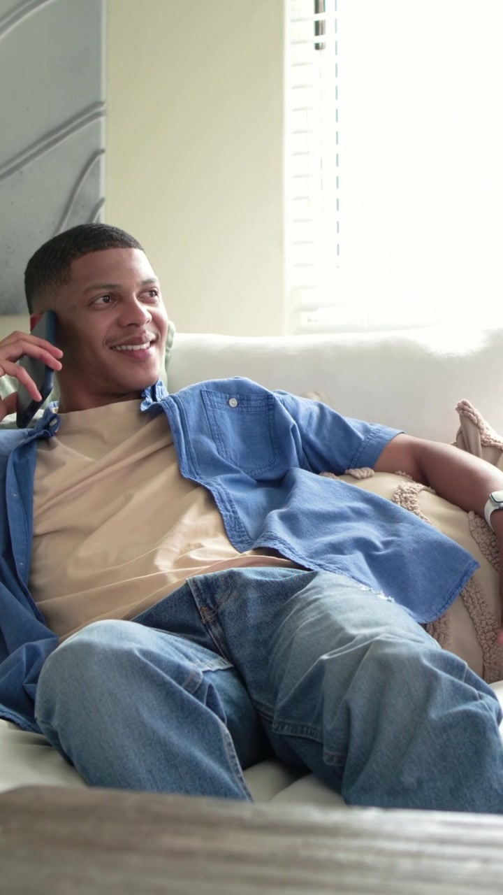 Video verticale: Uomo con camicia blu che scorre il telefono a casa sul divano, riceve una chiamata per una chiacchierata.