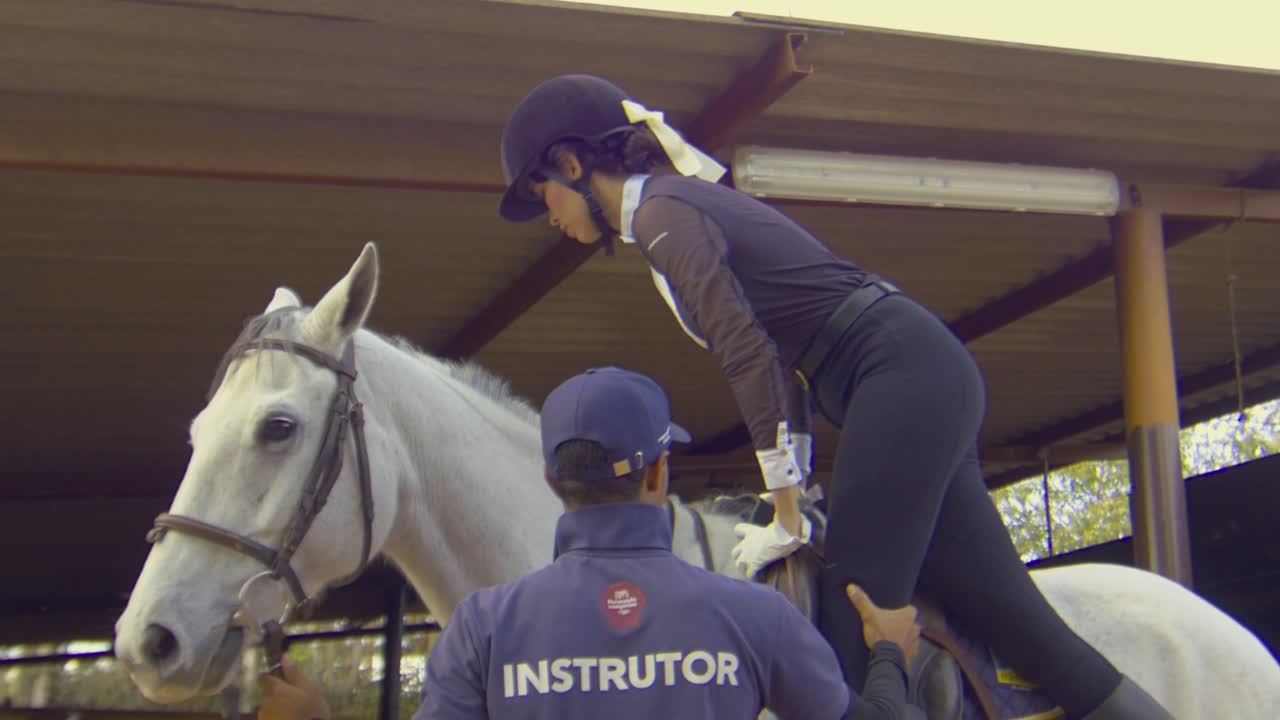 una toma cinematográfica en cámara lenta de una hermosa mujer subiendo a un caballo blanco mientras un instructor la está ayudando, slomo