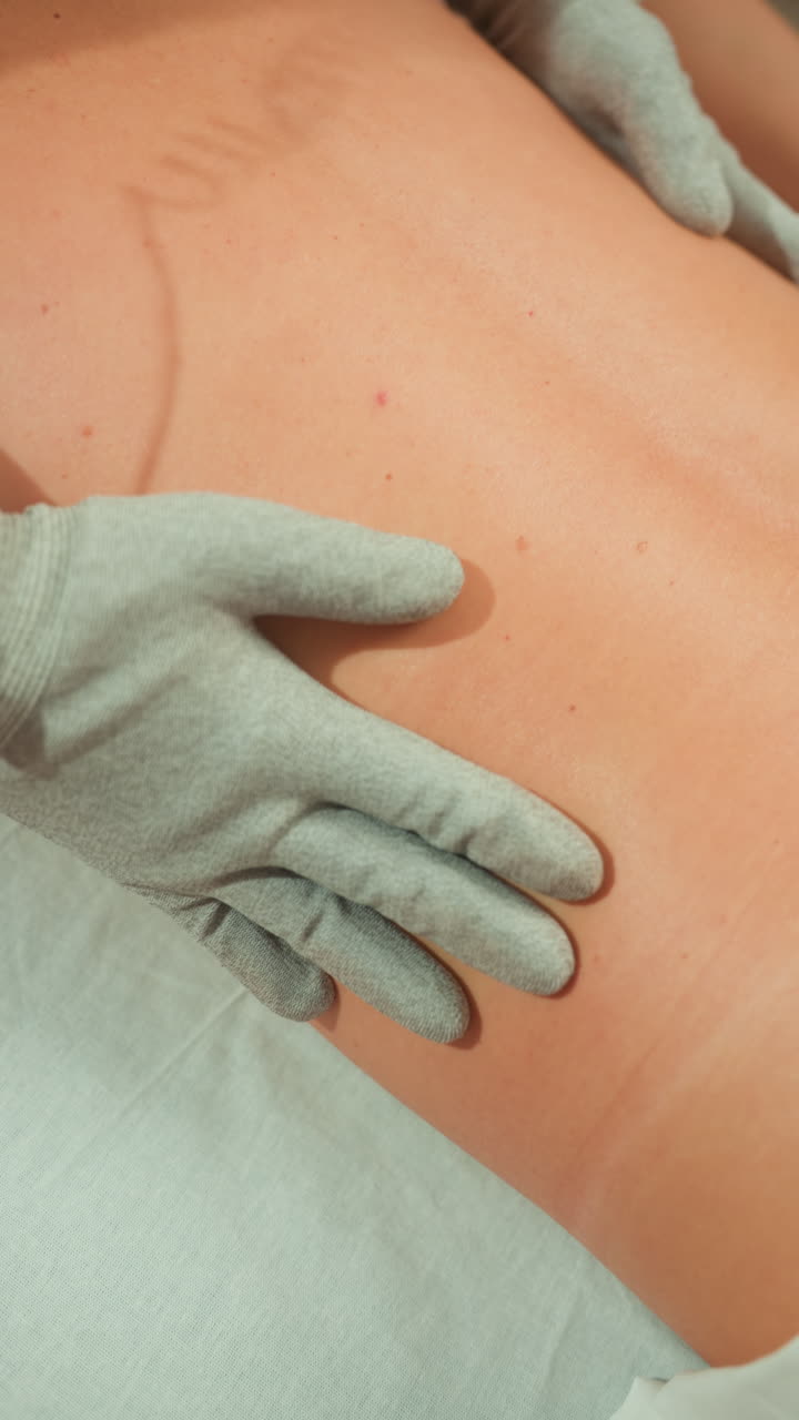 massaggio microcorrente con guanti sulla schiena, guanti conduttivi che erogano impulsi delicati sulla zona lombare scoperta in un ambiente spa clinico, tecnico esegue movimenti leviganti per alleviare il dolore, ridefinire i contorni e migliorare la pelle