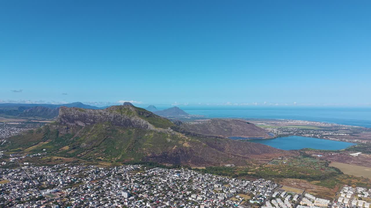 Expansive drone shot of Beau Bassin–Rose Hill, Mauritius, overlooking the coastline, ocean, mountains, and city below under a clear blue sky
