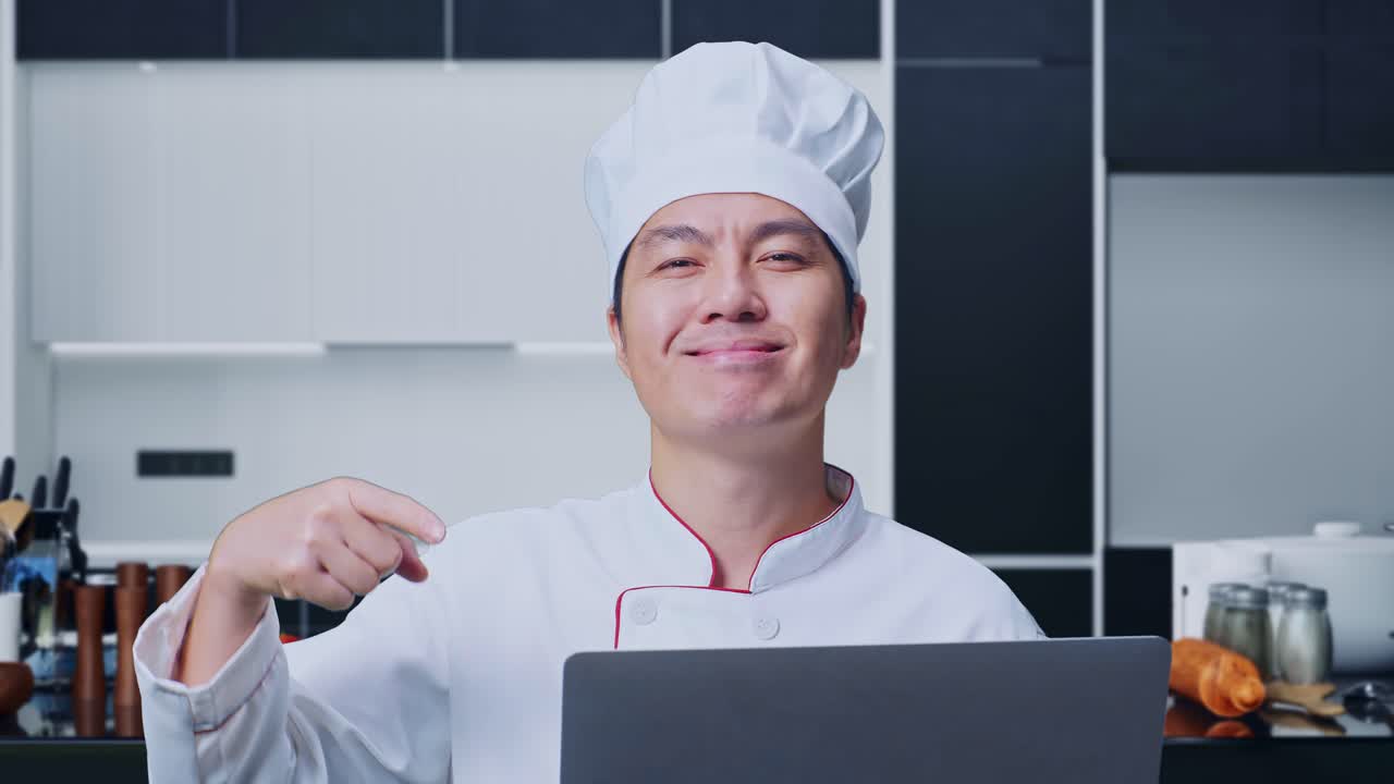 집 주방에 서 있는 동안 손에 노트북을 가리키며 미소 짓는 아시아인 요리사