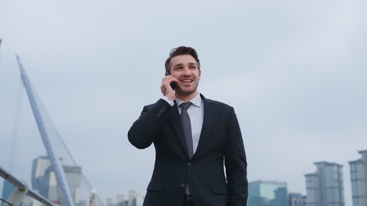 Plano em paralaxe, em câmara lenta e em ângulo baixo de um profissional de negócios positivo e feliz consultando um cliente em um smartphone enquanto está ao ar livre, tendo como plano de fundo edifícios altos de vidro modernos